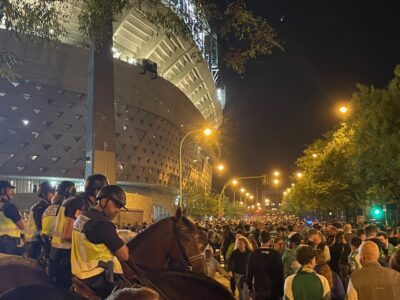 a large crowd outside a football stadium, watched over by police on horses.