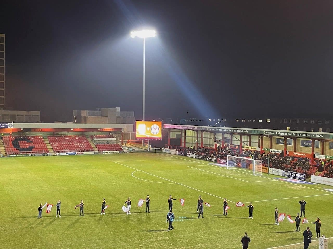 young people waving flags on a football pitch under floodlights before a game.