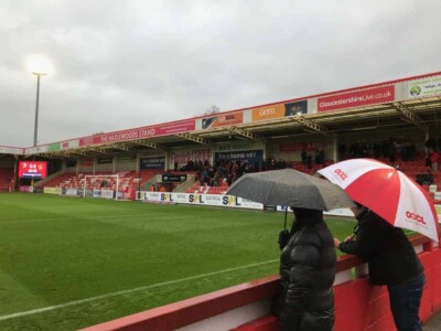 Supporters with umbrellas watching a game at Whaddon Road, home of Cheltenham Town FC.