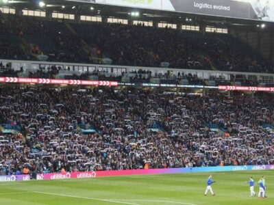football supporters holding up scarves in the stands while players warm up on the pitch.