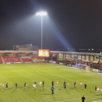 young people waving flags on a football pitch under floodlights before a game.