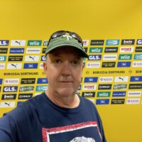 a man taking a selfie in front of stadium interview boards covered with logos